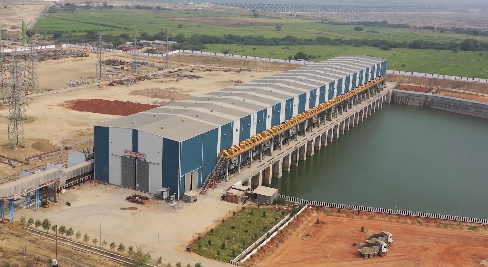 Aerial view of a long pump house building beside a fore-bay of water, with farmland behind